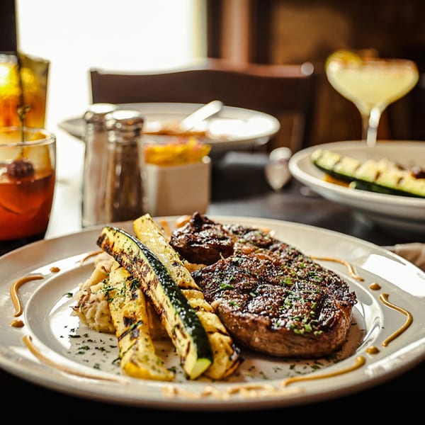 Grilled steak with mashed potatoes and zucchini, with drinks in the background at Silver Creek Beer Garden & Grille.