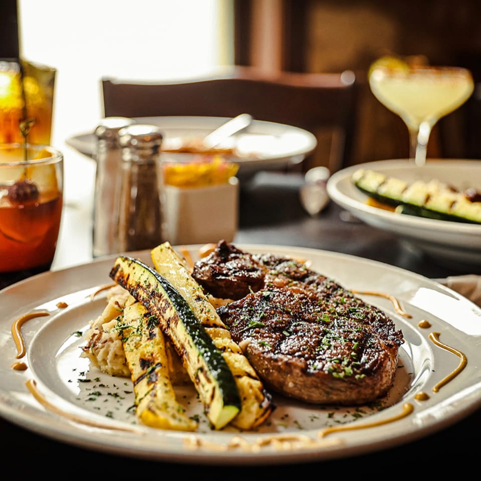 Grilled steak with mashed potatoes and zucchini, with drinks in the background at Silver Creek Beer Garden & Grille.