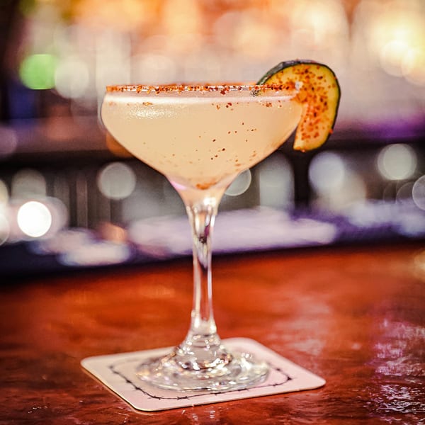 A light-colored cocktail in a coupe glass, garnished with a spiced rim and a cucumber slice.
