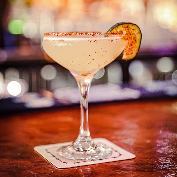 A light-colored cocktail in a coupe glass with a chili-salt rim and a cucumber slice.