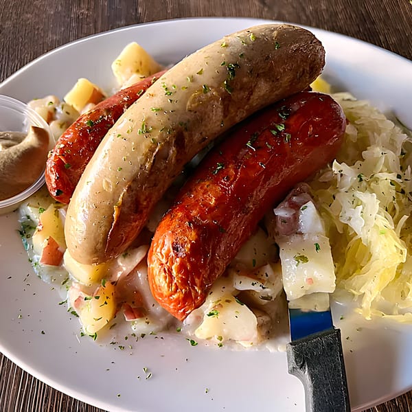 Two grilled sausages, one light and one dark, with potatoes, sauerkraut, and mustard on a white plate.