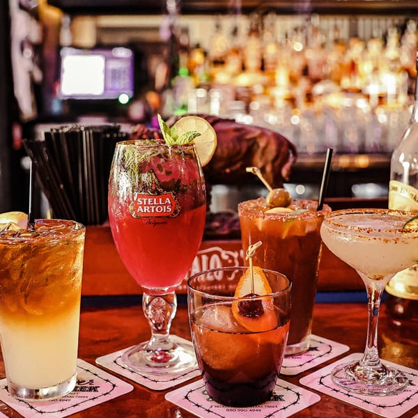 Five colorful cocktails and drinks on coasters on a bar, with bottles in the background.